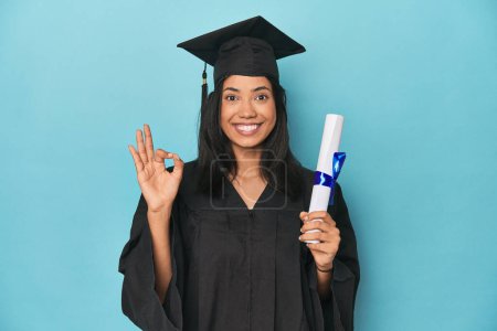 Photo for Filipina graduate with diploma on blue studio cheerful and confident showing ok gesture. - Royalty Free Image
