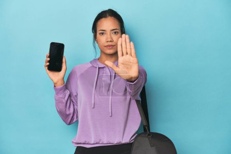 Photo for Filipina ready for gym with phone on blue standing with outstretched hand showing stop sign, preventing you. - Royalty Free Image