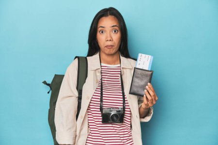 Photo for Filipina with camera, tickets, backpack on blue shrugs shoulders and open eyes confused. - Royalty Free Image