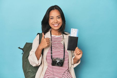 Photo for Filipina with camera, tickets, backpack on blue smiling and raising thumb up - Royalty Free Image