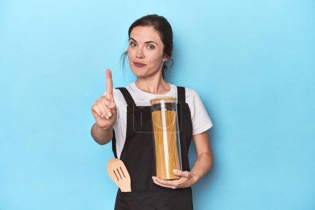 Téléchargez les photos : Chef femme avec pot de pâtes sur bleu montrant numéro un avec doigt. - en image libre de droit
