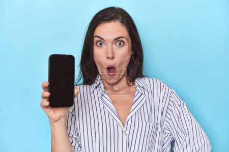 Téléchargez les photos : Femme surprise montrant écran de téléphone sur bleu - en image libre de droit