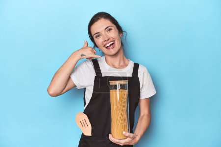 Téléchargez les photos : Chef femme avec pot de pâtes sur bleu montrant un geste d'appel téléphonique mobile avec les doigts. - en image libre de droit