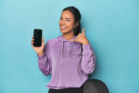 Téléchargez les photos : Filipina prêt pour la gym avec téléphone sur bleu montrant un geste d'appel de téléphone portable avec les doigts. - en image libre de droit