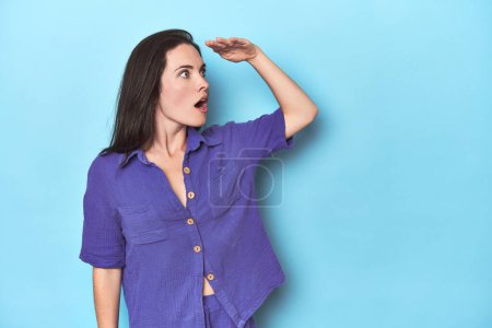 Photo for Young caucasian woman on blue backdrop looking far away keeping hand on forehead. - Royalty Free Image