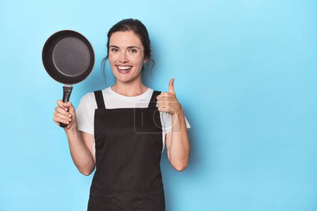 Photo for Woman with apron and pan on blue background smiling and raising thumb up - Royalty Free Image