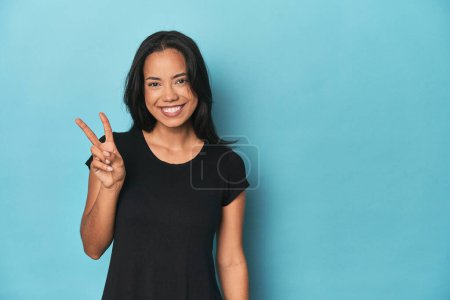 Foto de Filipina mujer joven en el estudio azul mostrando signo de victoria y sonriendo ampliamente. - Imagen libre de derechos