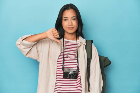 Téléchargez les photos : Femme philippine avec caméra et sac à dos sur bleu montrant un geste détestable, pouces vers le bas. Concept de désaccord. - en image libre de droit