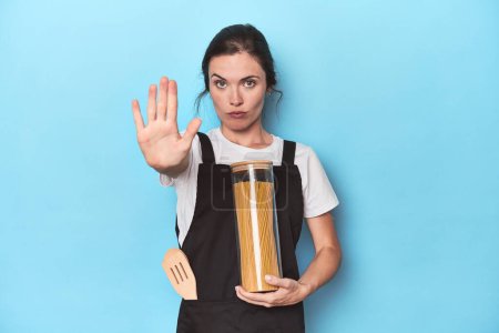 Téléchargez les photos : Chef femme avec pot de pâtes sur fond bleu debout avec la main tendue montrant stop signe, vous empêchant. - en image libre de droit