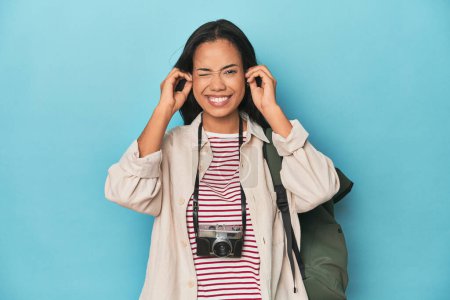 Téléchargez les photos : Philippine femme avec caméra et sac à dos sur bleu couvrant les oreilles avec les mains. - en image libre de droit