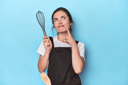 Téléchargez les photos : Chef dans tablier tenant fouet manuel, prêt à mélanger sur bleu - en image libre de droit