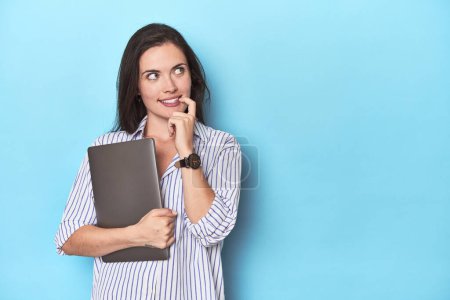 Téléchargez les photos : Jeune femme d'affaires tenant un ordinateur portable sur bleu détendu penser à quelque chose qui regarde un espace de copie. - en image libre de droit