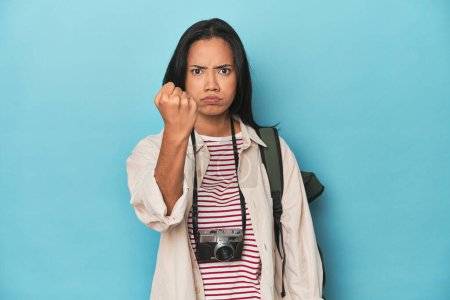Photo for Filipina woman with camera and backpack on blue showing fist to camera, aggressive facial expression. - Royalty Free Image