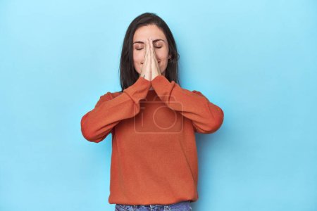 Téléchargez les photos : Jeune femme caucasienne sur fond bleu tenant la main dans la prière près de la bouche, se sent confiant. - en image libre de droit