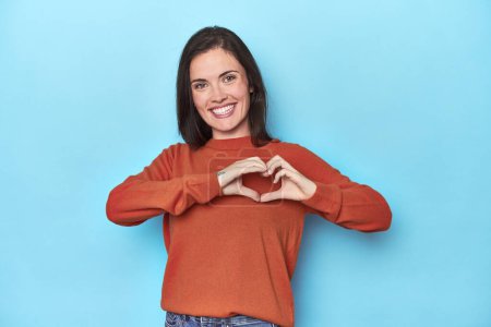 Photo for Young caucasian woman on blue backdrop smiling and showing a heart shape with hands. - Royalty Free Image