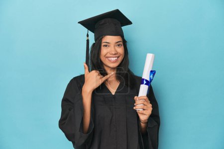 Photo for Filipina graduate with diploma on blue studio showing a mobile phone call gesture with fingers. - Royalty Free Image