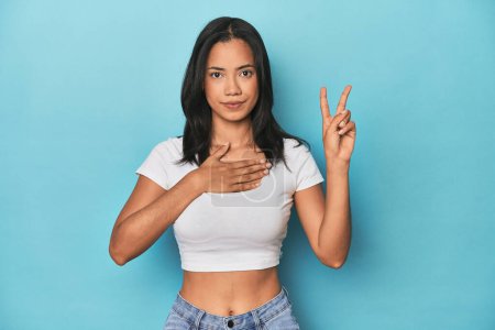 Téléchargez les photos : Philippine jeune femme sur studio bleu prendre un serment, mettre la main sur la poitrine. - en image libre de droit