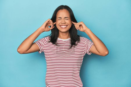 Photo for Filipina young woman on blue studio covering ears with fingers, stressed and desperate by a loudly ambient. - Royalty Free Image