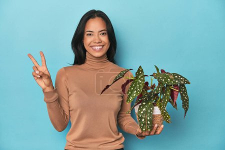 Photo for Filipina holding a plant on blue studio showing number two with fingers. - Royalty Free Image