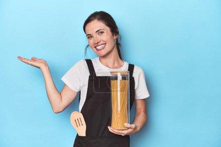 Foto de Chef mujer con tarro de pasta en azul mostrando un espacio de copia en una palma y sosteniendo otra mano en la cintura. - Imagen libre de derechos