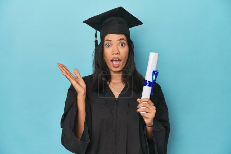 Photo for Filipina graduate with diploma on blue studio surprised and shocked. - Royalty Free Image