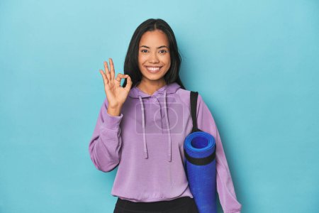 Foto de Atleta filipina con esterilla de yoga en azul alegre y confiado mostrando buen gesto. - Imagen libre de derechos