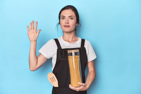 Foto de Chef mujer con tarro de pasta en azul sonriente alegre mostrando el número cinco con los dedos. - Imagen libre de derechos