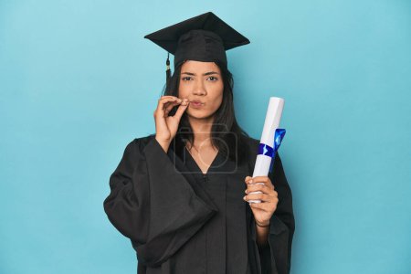 Photo for Filipina graduate with diploma on blue studio with fingers on lips keeping a secret. - Royalty Free Image