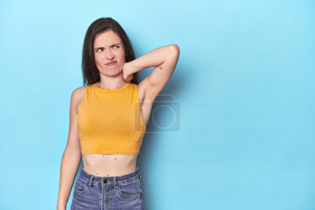 Photo for Young caucasian woman on blue backdrop touching back of head, thinking and making a choice. - Royalty Free Image