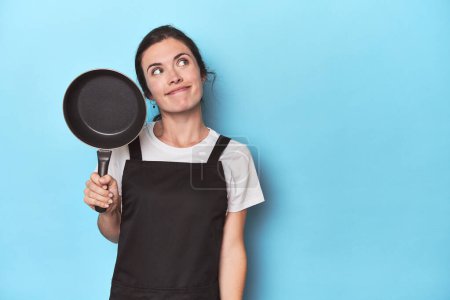 Photo for Woman with apron and pan on blue background dreaming of achieving goals and purposes - Royalty Free Image