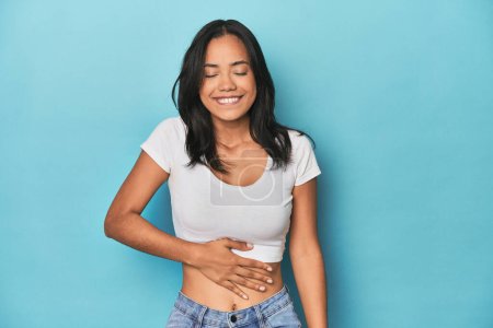 Photo for Filipina young woman on blue studio touches tummy, smiles gently, eating and satisfaction concept. - Royalty Free Image