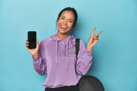 Photo for Filipina ready for gym with phone on blue joyful and carefree showing a peace symbol with fingers. - Royalty Free Image
