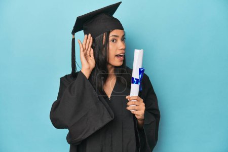 Téléchargez les photos : Filipina diplômé avec diplôme sur studio bleu en essayant d'écouter un potins. - en image libre de droit