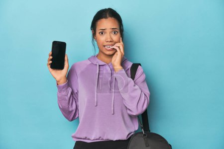 Photo for Filipina ready for gym with phone on blue biting fingernails, nervous and very anxious. - Royalty Free Image