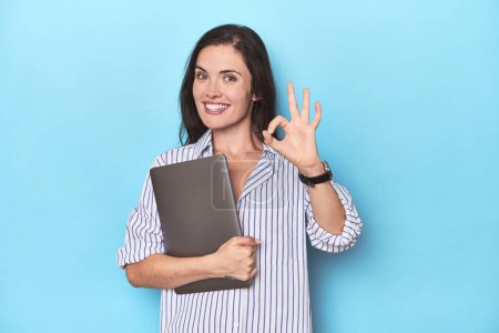 Photo for Young businesswoman holding laptop on blue cheerful and confident showing ok gesture. - Royalty Free Image