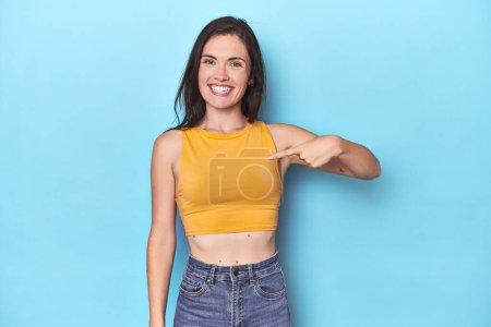Téléchargez les photos : Jeune femme caucasienne sur fond bleu personne pointant à la main vers un espace de copie de chemise, fière et confiante - en image libre de droit