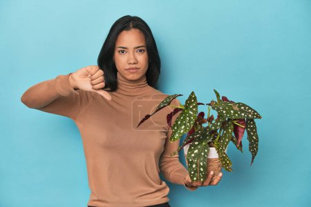 Foto de Filipina sosteniendo una planta en el estudio azul mostrando un gesto de aversión, pulgares hacia abajo. Concepto de desacuerdo. - Imagen libre de derechos