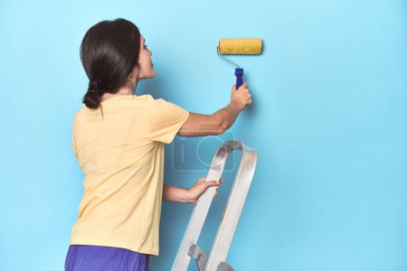 Téléchargez les photos : Femme peignant un mur bleu d'une échelle - en image libre de droit