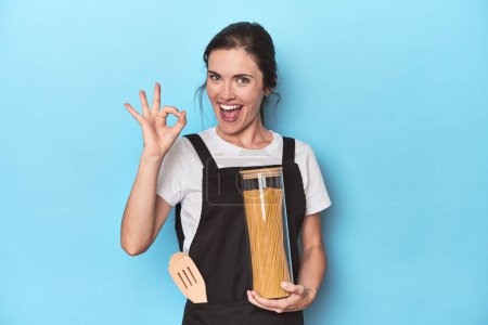 Photo for Chef woman with pasta jar on blue cheerful and confident showing ok gesture. - Royalty Free Image