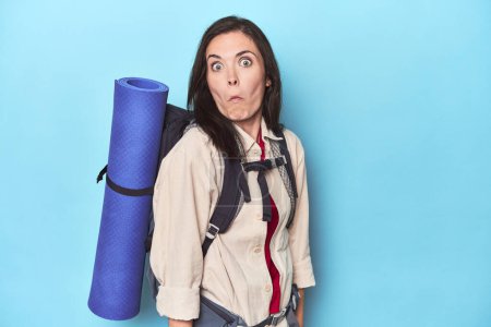 Téléchargez les photos : Femme aventureuse avec sac à dos sur les épaules bleues haussant les épaules et les yeux ouverts confus. - en image libre de droit