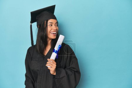 Photo for Filipina graduate with diploma on blue studio points with thumb finger away, laughing and carefree. - Royalty Free Image