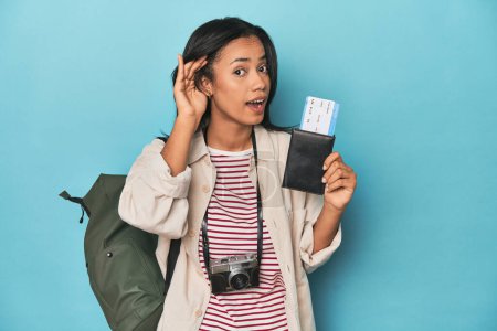 Téléchargez les photos : Filipina avec caméra, billets, sac à dos sur bleu en essayant d'écouter un potins. - en image libre de droit