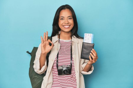 Téléchargez les photos : Filipina avec caméra, billets, sac à dos sur bleu gai et confiant montrant geste ok. - en image libre de droit