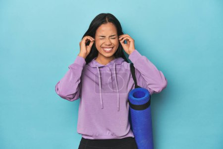 Photo for Filipina athlete with yoga mat on blue covering ears with hands. - Royalty Free Image
