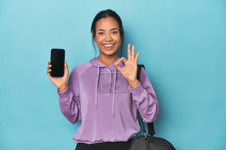 Photo for Filipina ready for gym with phone on blue cheerful and confident showing ok gesture. - Royalty Free Image