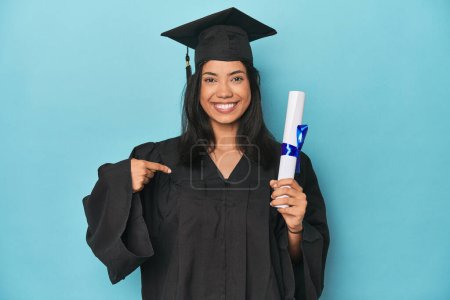 Photo for Filipina graduate with diploma on blue studio person pointing by hand to a shirt copy space, proud and confident - Royalty Free Image