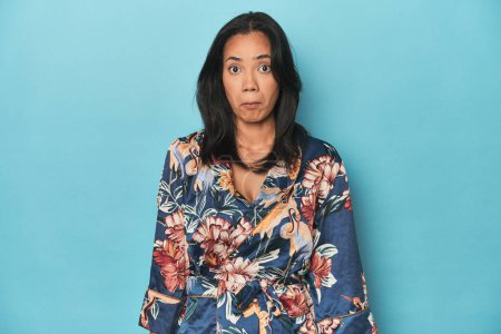 Photo for Filipina in floral kimono on blue studio shrugs shoulders and open eyes confused. - Royalty Free Image
