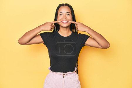Photo for Young Indonesian woman on yellow studio backdrop smiles, pointing fingers at mouth. - Royalty Free Image