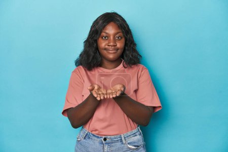 Téléchargez les photos : Jeune femme courbée afro-américaine tenant quelque chose avec des paumes, offrant à la caméra. - en image libre de droit