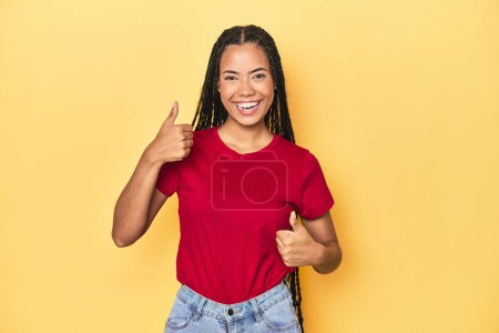 Photo for Young Indonesian woman on yellow studio backdrop raising both thumbs up, smiling and confident. - Royalty Free Image